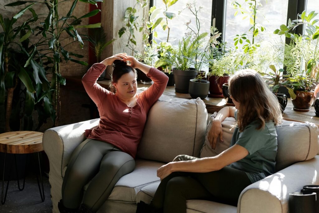 Two women enjoying a relaxed conversation on a sofa in a bright room filled with plants.
