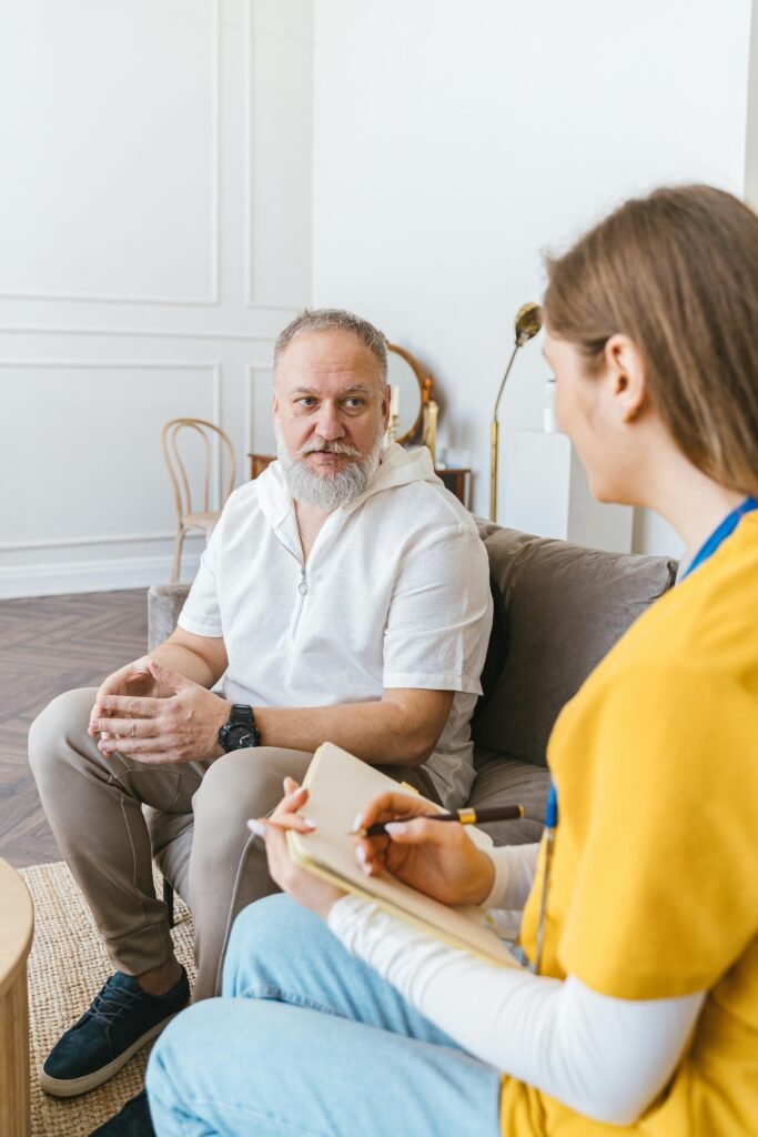 A senior man in a white shirt talks to a woman volunteer taking notes indoors.